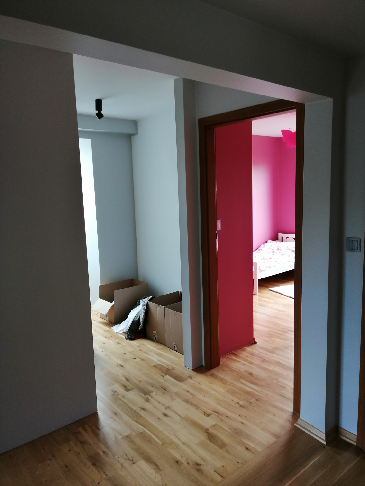 Widok przez dwa otwory drzwiowe na pokój z drewnianą podłogą, kartonami i różowy pokój dziecięcy z łóżkiem. Nowoczesne oświetlenie sufitowe w pierwszym pokoju.