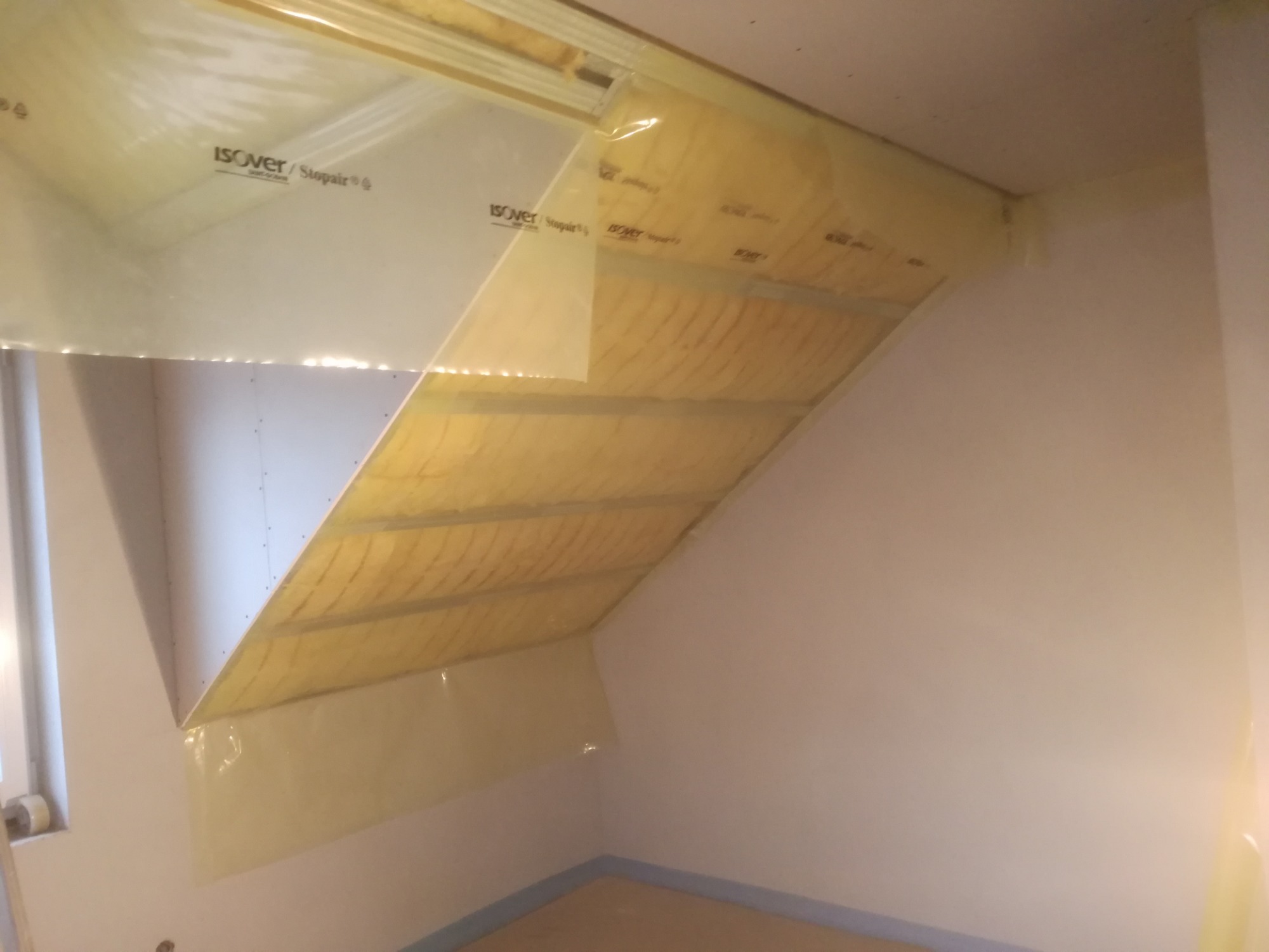 Izolacja poddasza wełną mineralną, zabezpieczona folią paroizolacyjną z widocznym logo Isover, częściowo zakryta białą płytą. Widoczne mocowania płyty do konstrukcji.