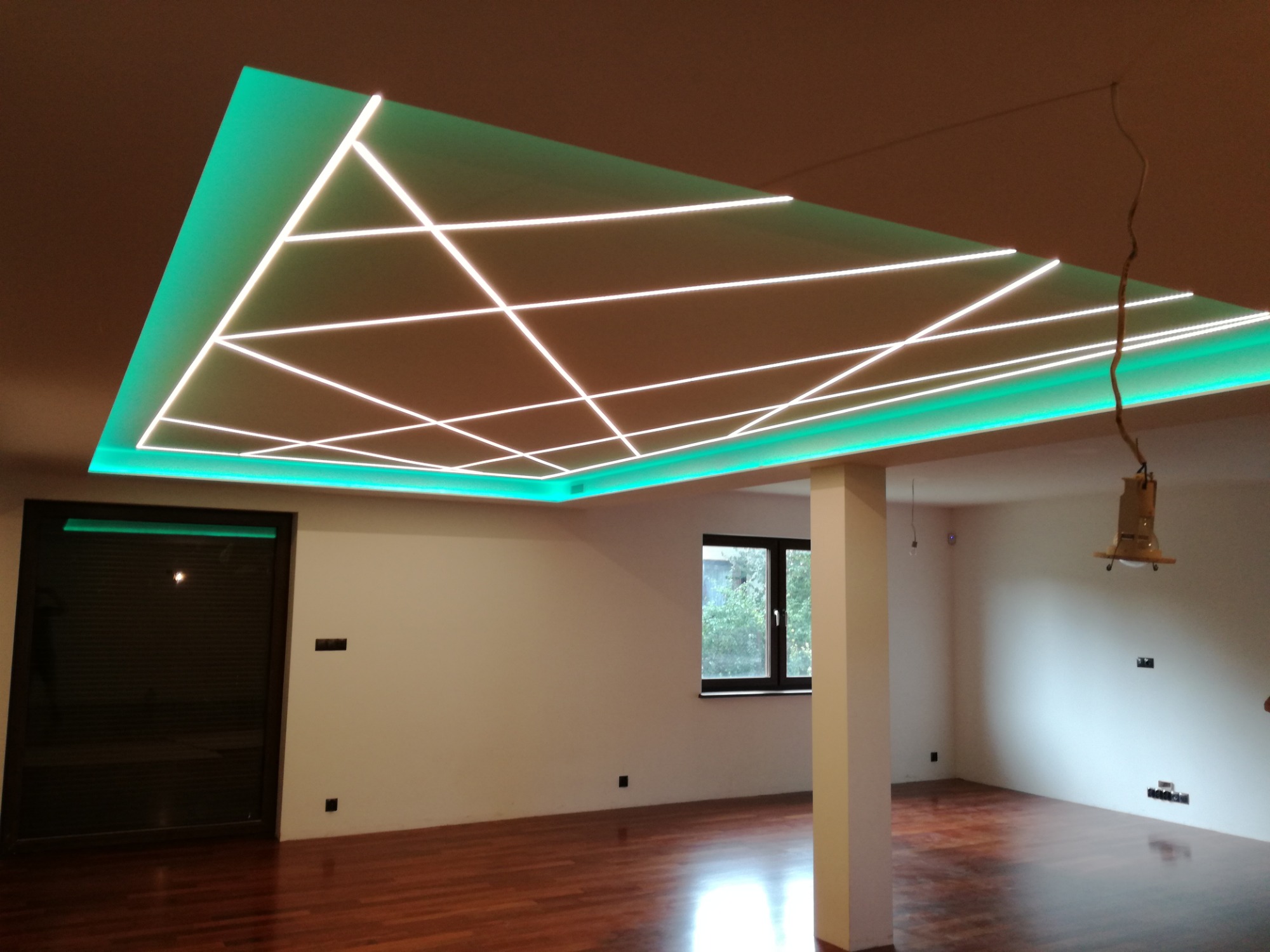 Nowoczesne wnętrze z podwieszanym sufitem z zielonym podświetleniem LED i geometrycznym wzorem z białych linii świetlnych, parkiet na podłodze, widok na okno i drzwi.