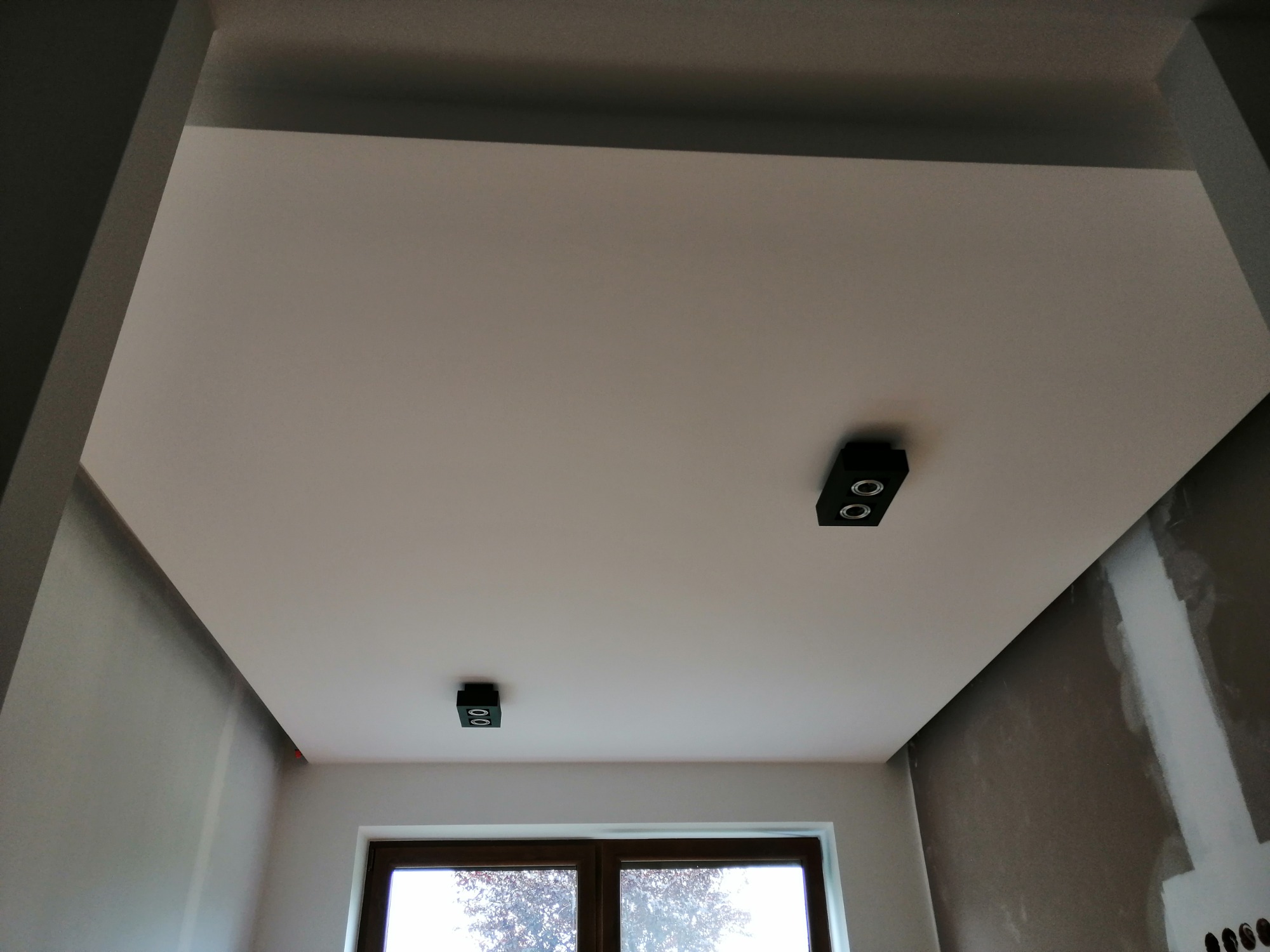 Pomieszczenie po malowaniu sufitu na jasny kolor z zamontowanymi dwoma czarnymi lampami punktowymi. Ściany częściowo przygotowane do dalszych prac wykończeniowych, widoczne ślady po szpachlowaniu.