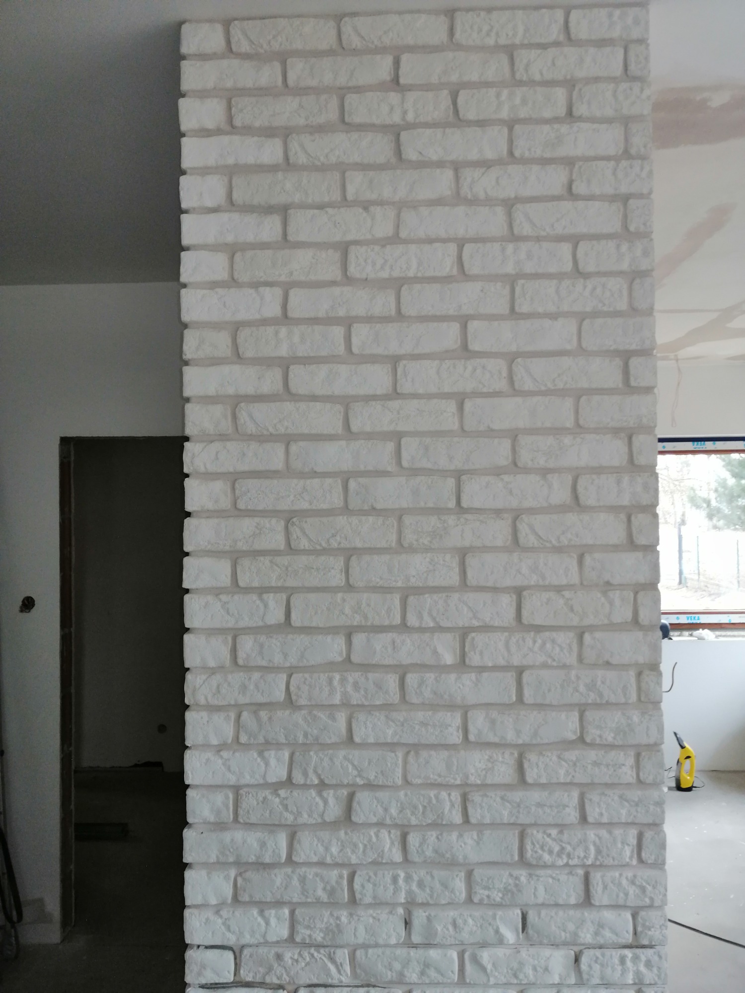 Fragment ściany wykończonej białymi płytkami imitującymi cegłę, widoczny w trakcie remontu wnętrza, z fragmentem niezakończonej gładzi na suficie i oknem w tle.