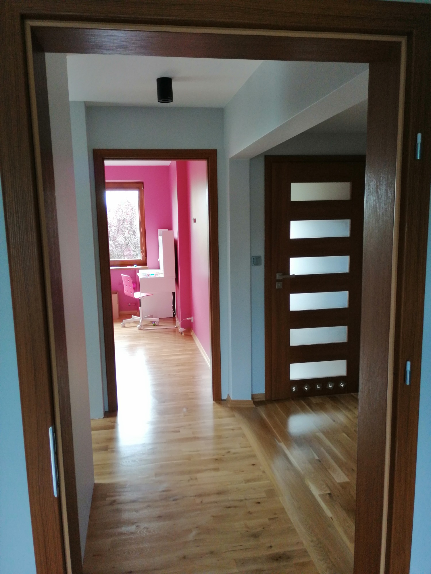 Widok na korytarz z drewnianą podłogą i trzema parami drzwi w Zduńskiej Woli. Jedne drzwi z przeszkleniami, drugie prowadzą do różowego pokoju z biurkiem.