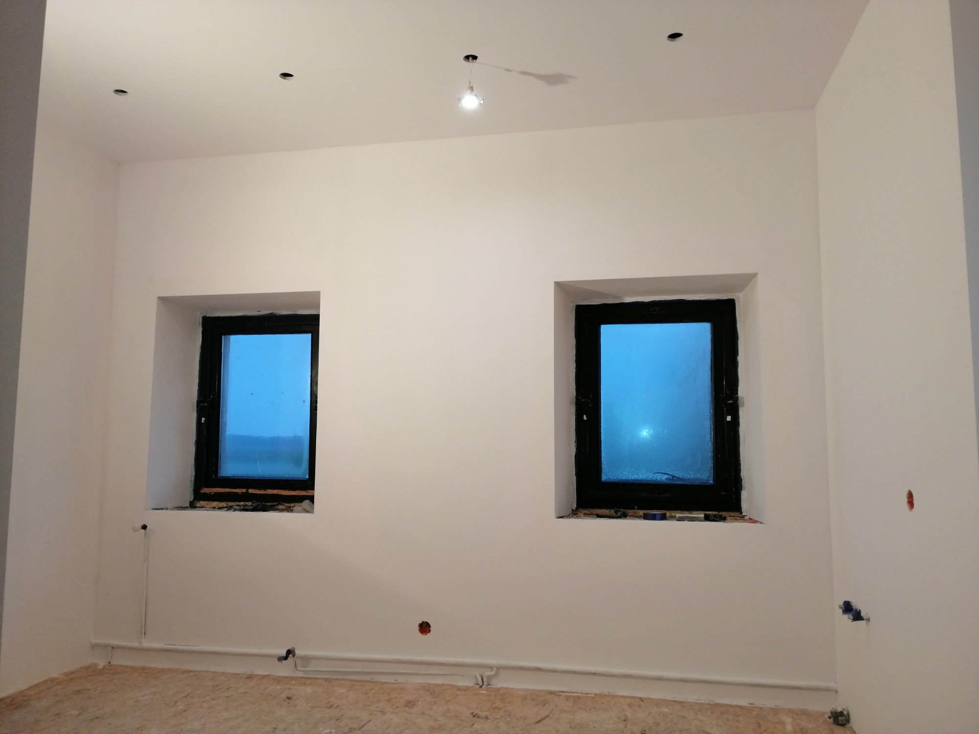 Wnętrze pokoju po malowaniu ścian na biało, widoczne dwa okna z czarnymi ramami, instalacja elektryczna w ścianach i rury grzewcze przy podłodze, surowa podłoga przygotowana do dalszych prac...