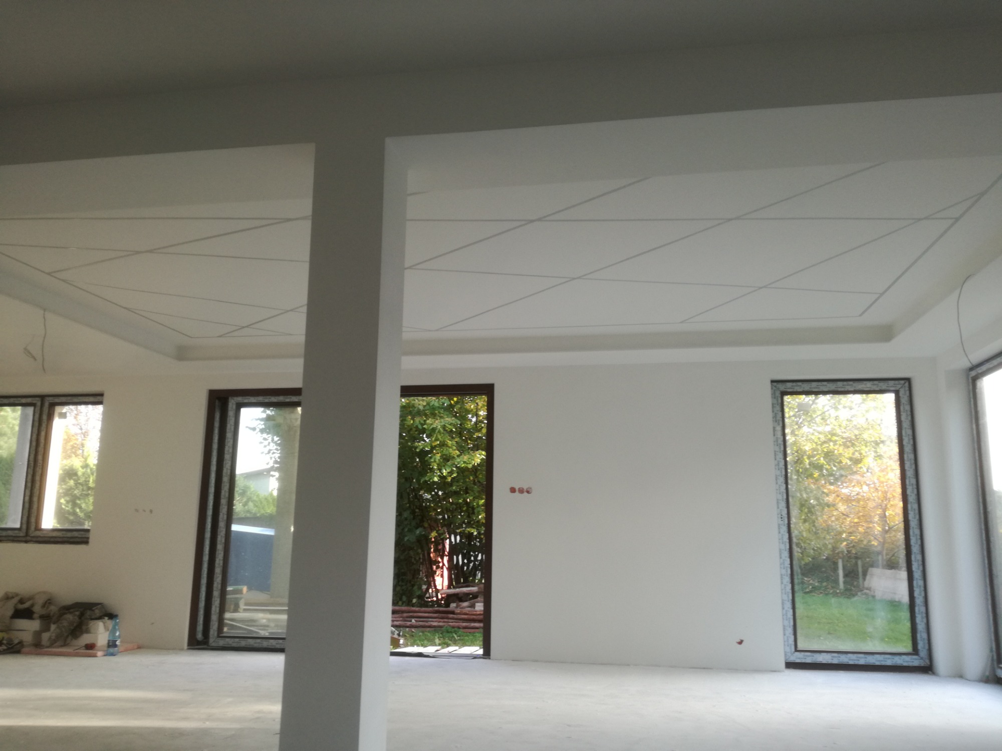 Surowe wnętrze z widocznymi elementami wykończeniowymi sufitu, w tym geometryczny wzór na podwieszanym suficie, duże okna z widokiem na zieleń i surowa, szara podłoga.