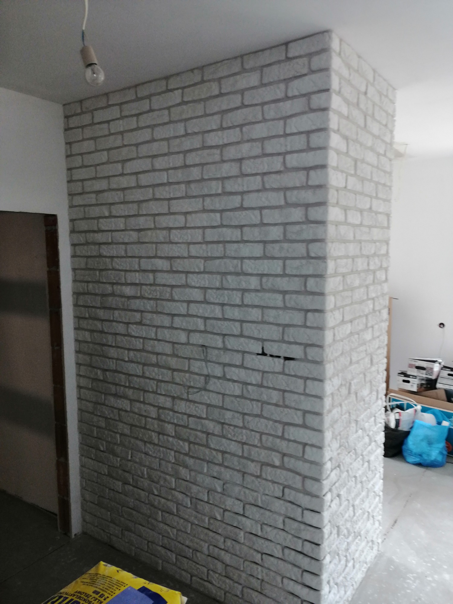 Ściana wykończona dekoracyjną cegłą w odcieniach szarości, tworząca narożnik w pomieszczeniu w trakcie prac wykończeniowych, widoczne niedoskonałości fugowania i surowa żarówka na kablu.