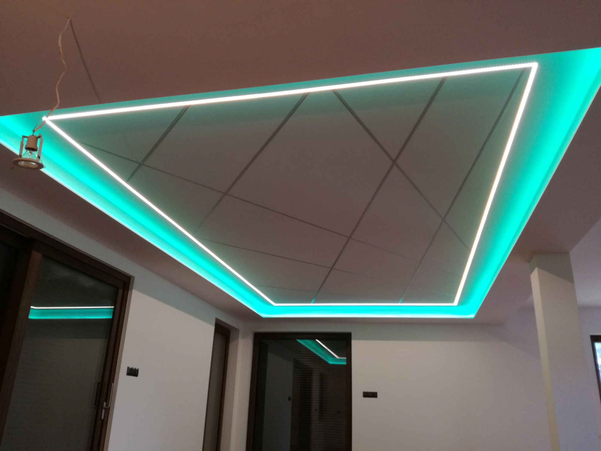 Podwieszany sufit z płyt z podświetleniem LED w kształcie trójkąta, widoczne linie podziału płyt i wisząca lampa z kablem.