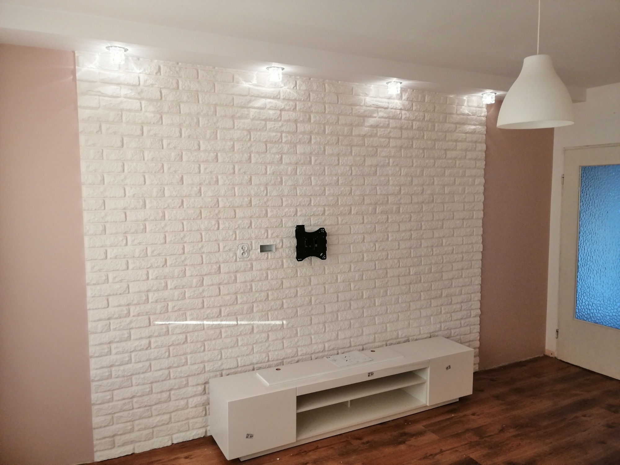 Wykończona ściana z białej cegły dekoracyjnej z zamontowanym uchwytem do telewizora, oświetlona punktowymi lampkami sufitowymi i wiszącą lampą, z białą szafką RTV na podłodze z paneli.