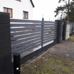 Nowoczesne ogrodzenie posesji z szarymi, poziomymi przęsłami i czarnymi, betonowymi słupkami oraz podmurówką, na tle domu z numerem 93.
