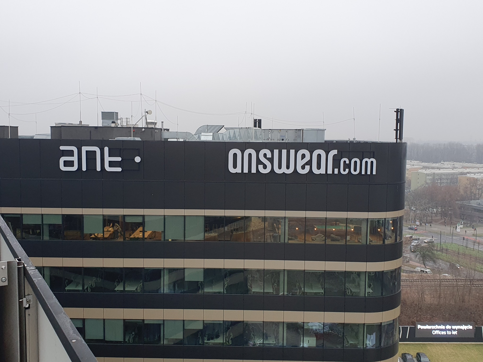 Budynek biurowy z logo 'ant.' i 'answear.com' na dachu, widok z góry na miasto w pochmurny dzień.