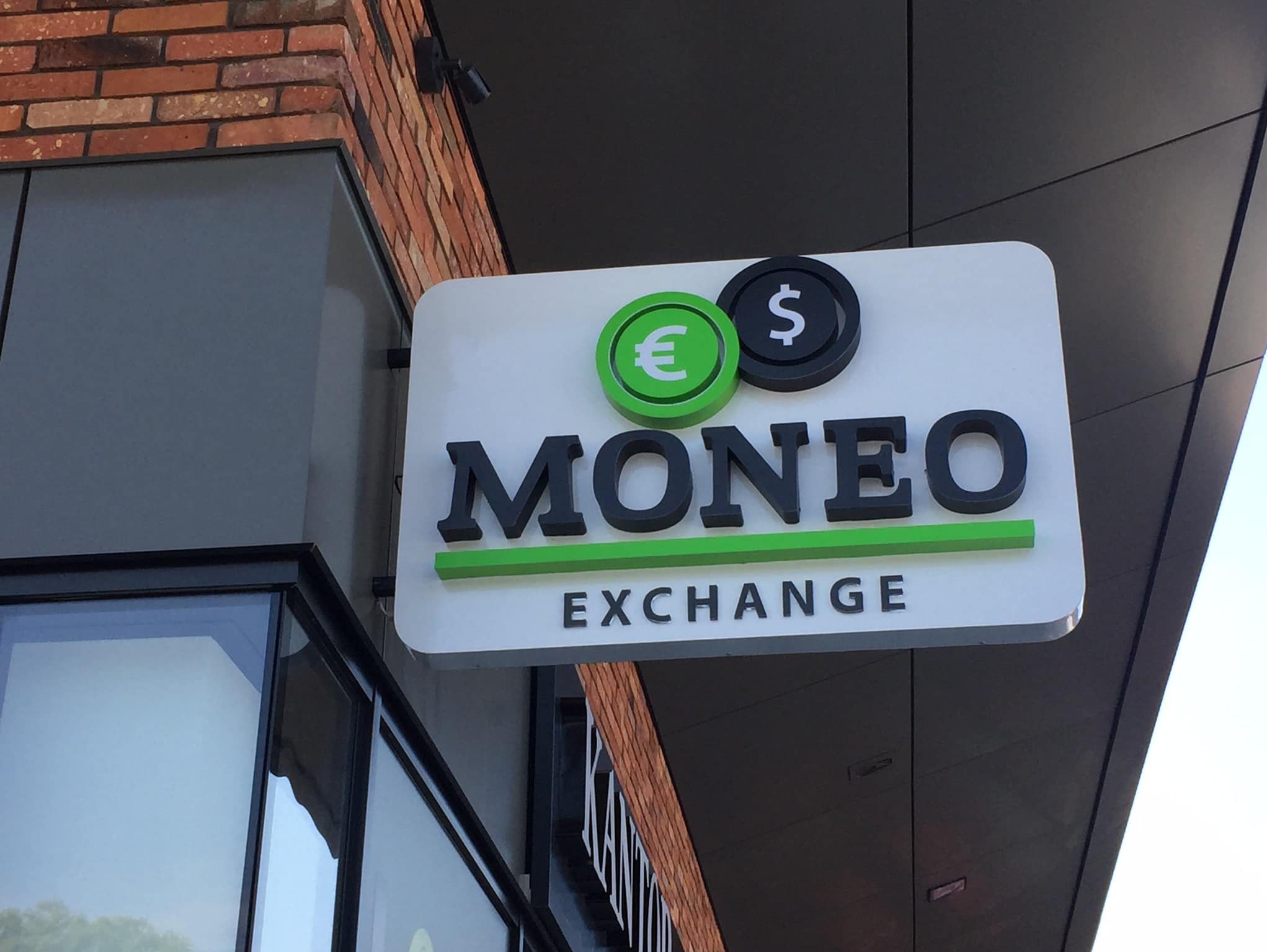 Kaseton reklamowy z napisem MONEO EXCHANGE, z logo przedstawiającym zielone euro i czarne dolary, zamontowany na elewacji budynku.