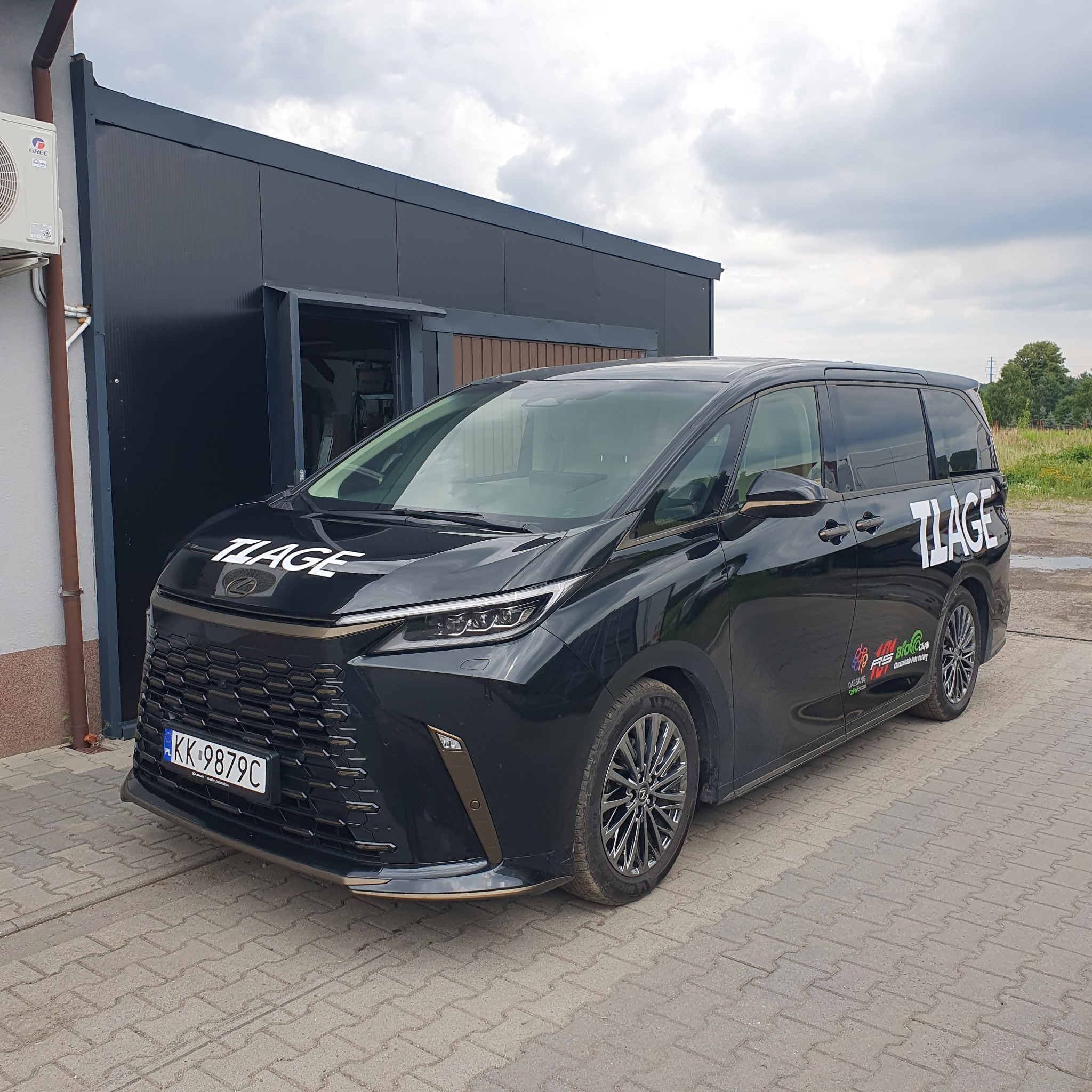 Czarny minivan z oklejonym logo 'TLAGE' na masce i boku, zaparkowany przed szarym budynkiem z klimatyzatorem na ścianie.