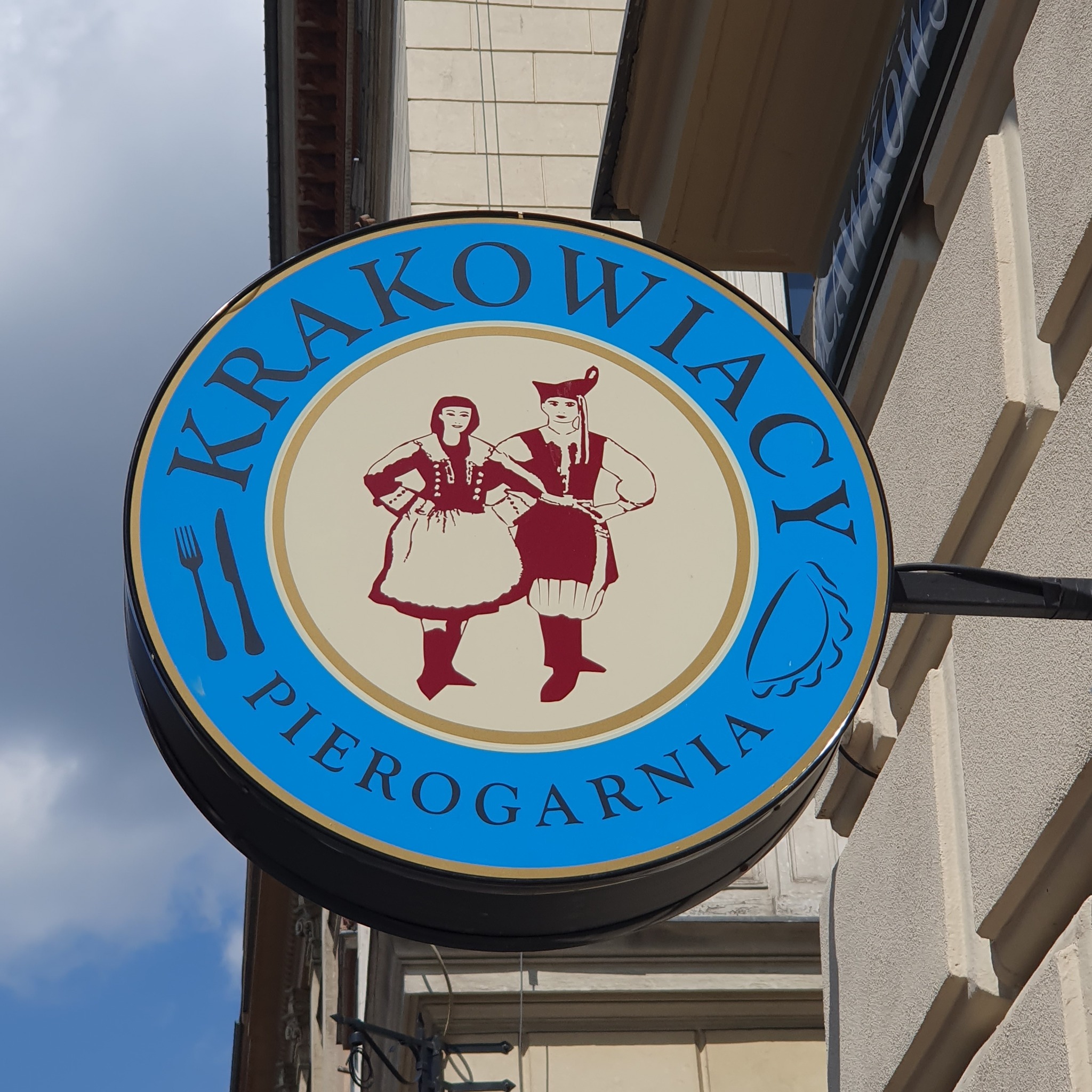 Okrągły szyld z nazwą 'KRAKOWIACY PIEROGARNIA', z ilustracją pary w strojach ludowych, stylizowany na retro, z widelcem i pierogiem po bokach.