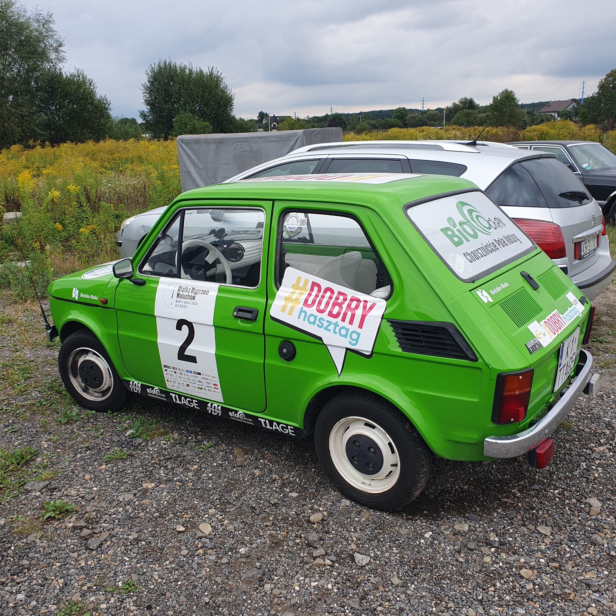 Zielony Fiat 126p z numerem startowym 2 i naklejkami sponsorskimi, w tym hasłem #DOBRYhasztag, zaparkowany na żwirowym parkingu.