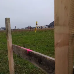 Tyczenie działki: widok przez drewniany płot z różową farbą na desce, w tle żółty tachimetr na zielonym polu i zabudowa jednorodzinna w oddali.