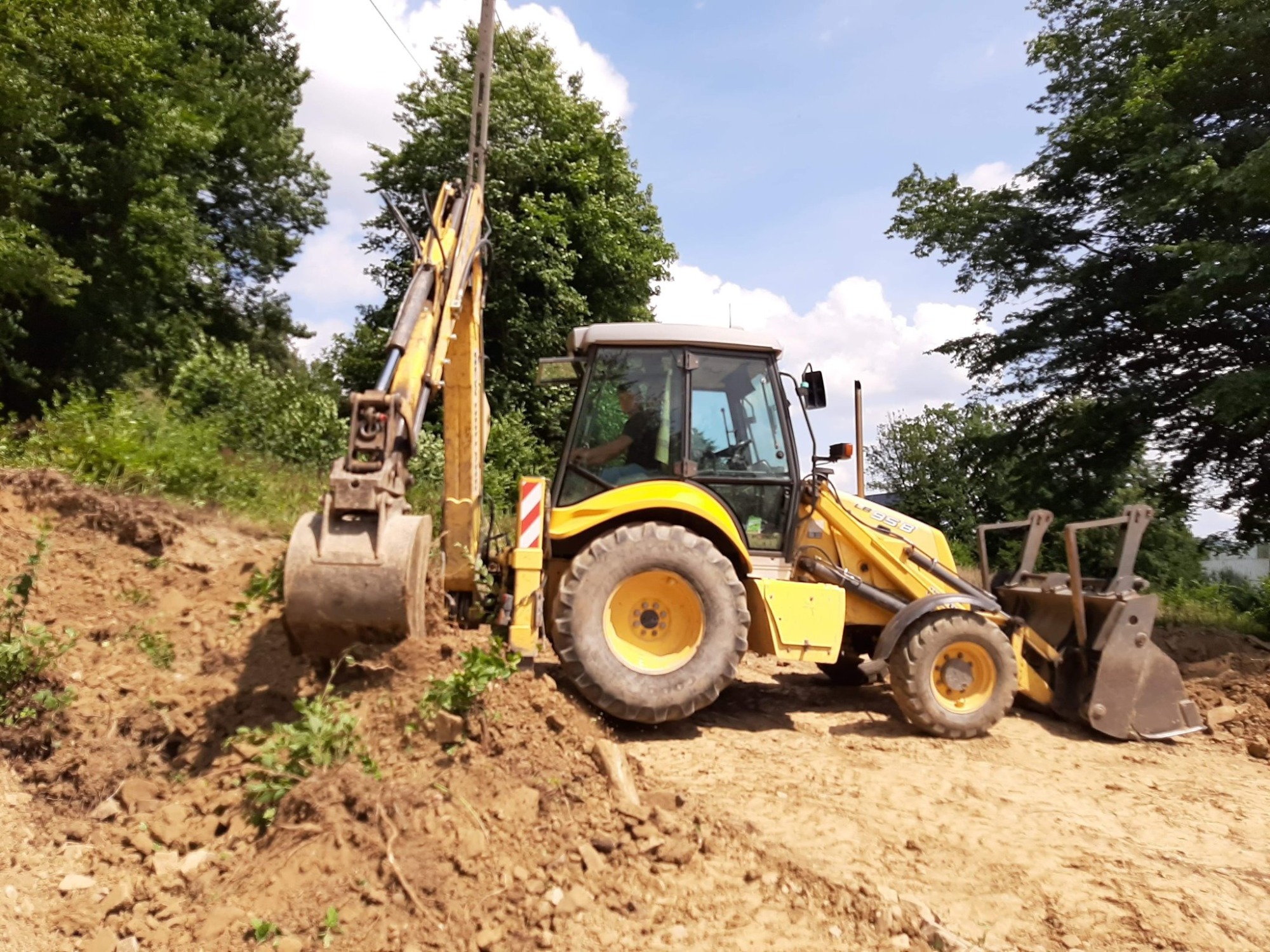 Żółta koparko-ładowarka na nierównym, ziemnym terenie, z widocznymi śladami opon i łyżką zagłębiającą się w ziemię, operator w kabinie, drzewa w tle.