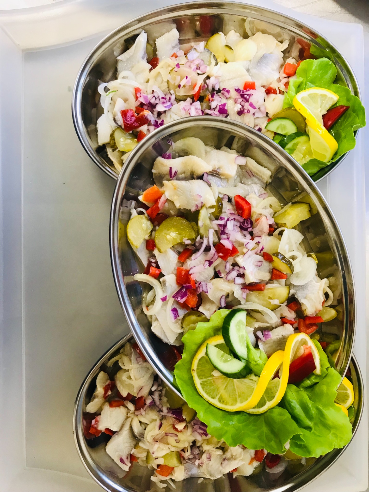 Trzy metalowe miski z sałatką śledziową udekorowaną czerwoną papryką, czerwoną cebulą, ogórkami kiszonymi oraz cytryną i sałatą na białej tacy.