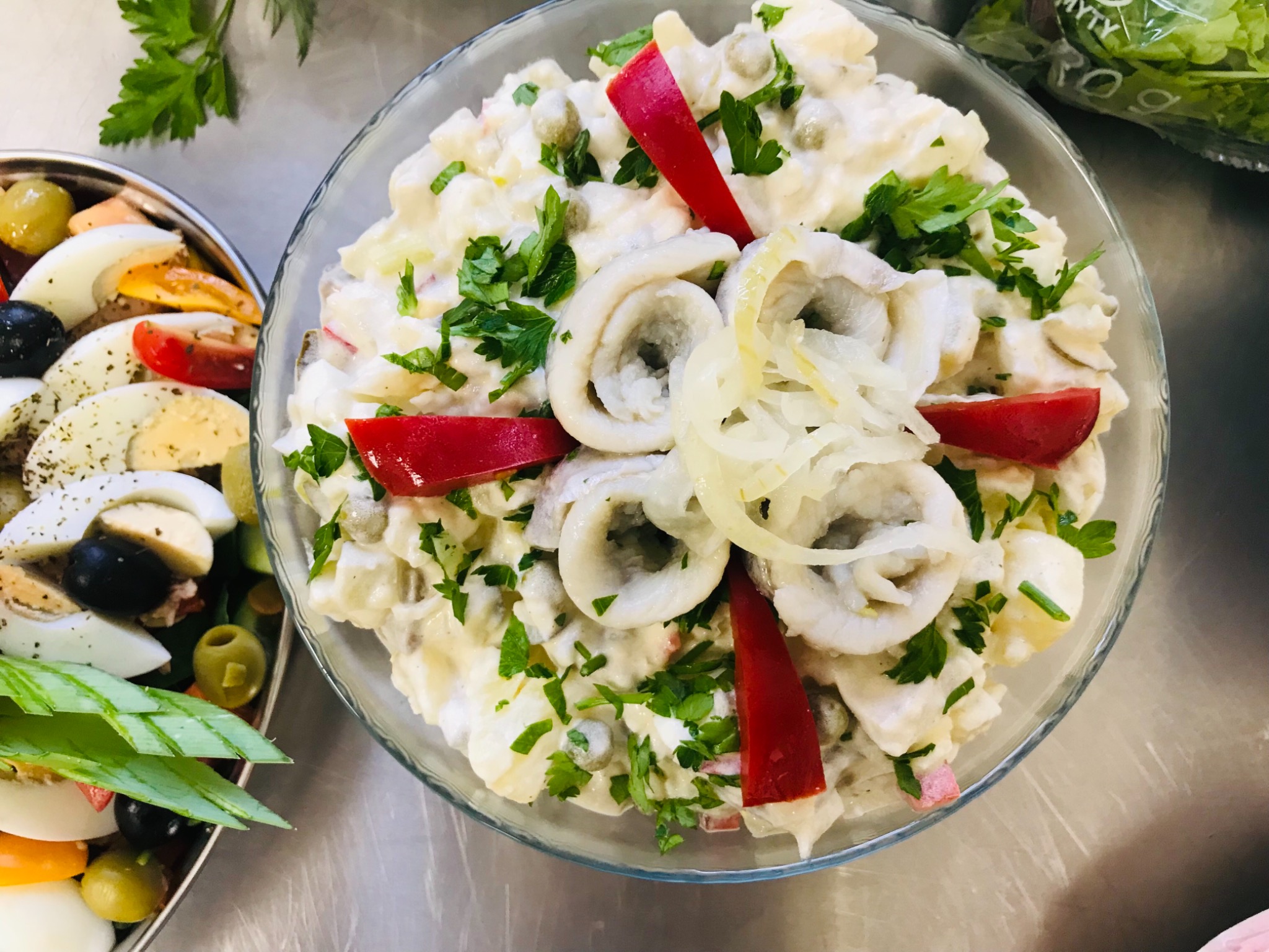 Sałatka jarzynowa z pierścieniami kalmarów i papryką, podana w szklanej misce, obok taca z jajkami, oliwkami i zielonym szczypiorkiem.