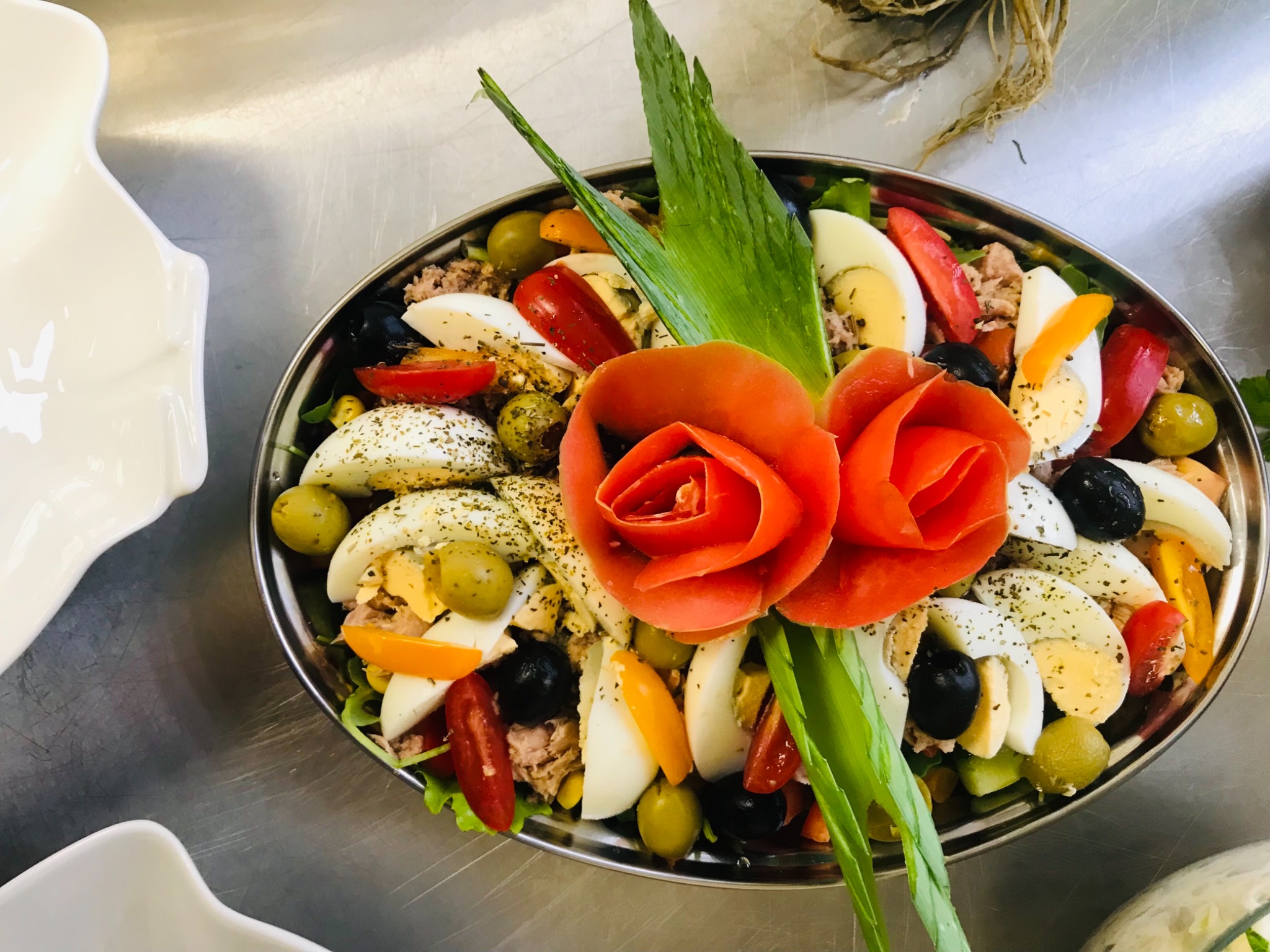 Dekoracyjna sałatka z tuńczykiem, jajkiem, oliwkami i pomidorami, ułożona na metalowej tacy z centralną dekoracją z uformowanych pomidorów przypominających róże i zielonych liści.
