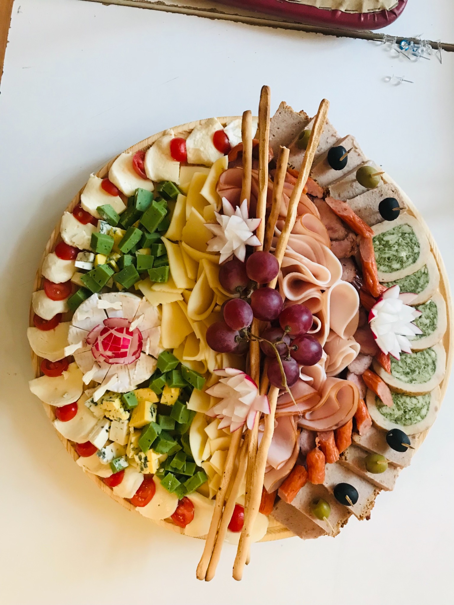 Dekoracyjna drewniana deska z selekcją serów (w tym kostki zielonego sera), wędlin, winogron, krakersów i rzodkiewek, ułożonych w segmenty.