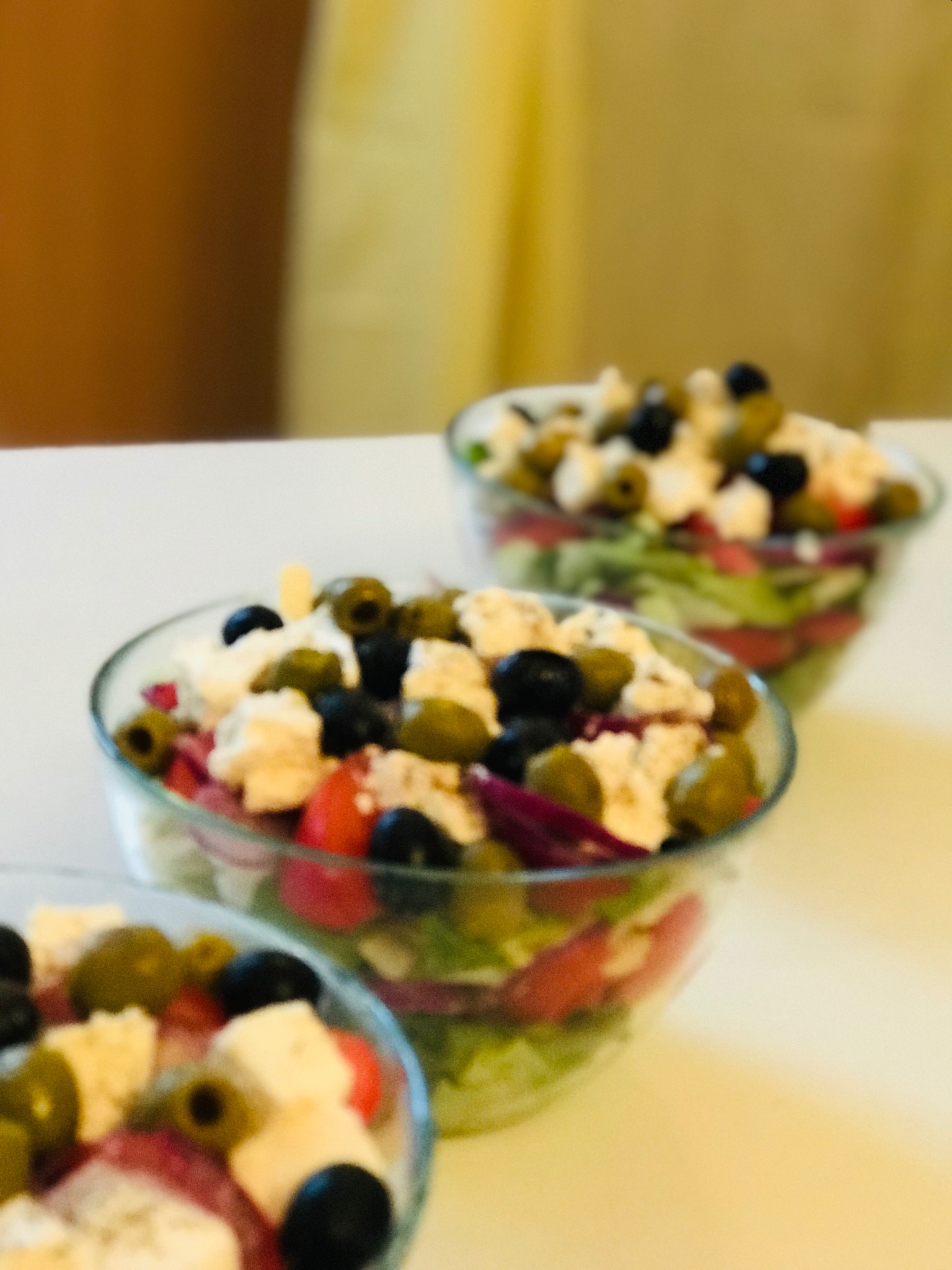 Trzy szklane miseczki z sałatką grecką, z widocznymi oliwkami, serem feta, pomidorami i czerwoną cebulą, ustawione w rzędzie w Krzeszowicach.