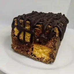 Kwadratowy kawałek ciasta z polewą czekoladową na białym talerzu, widoczne warstwy jasnego i ciemnego ciasta.