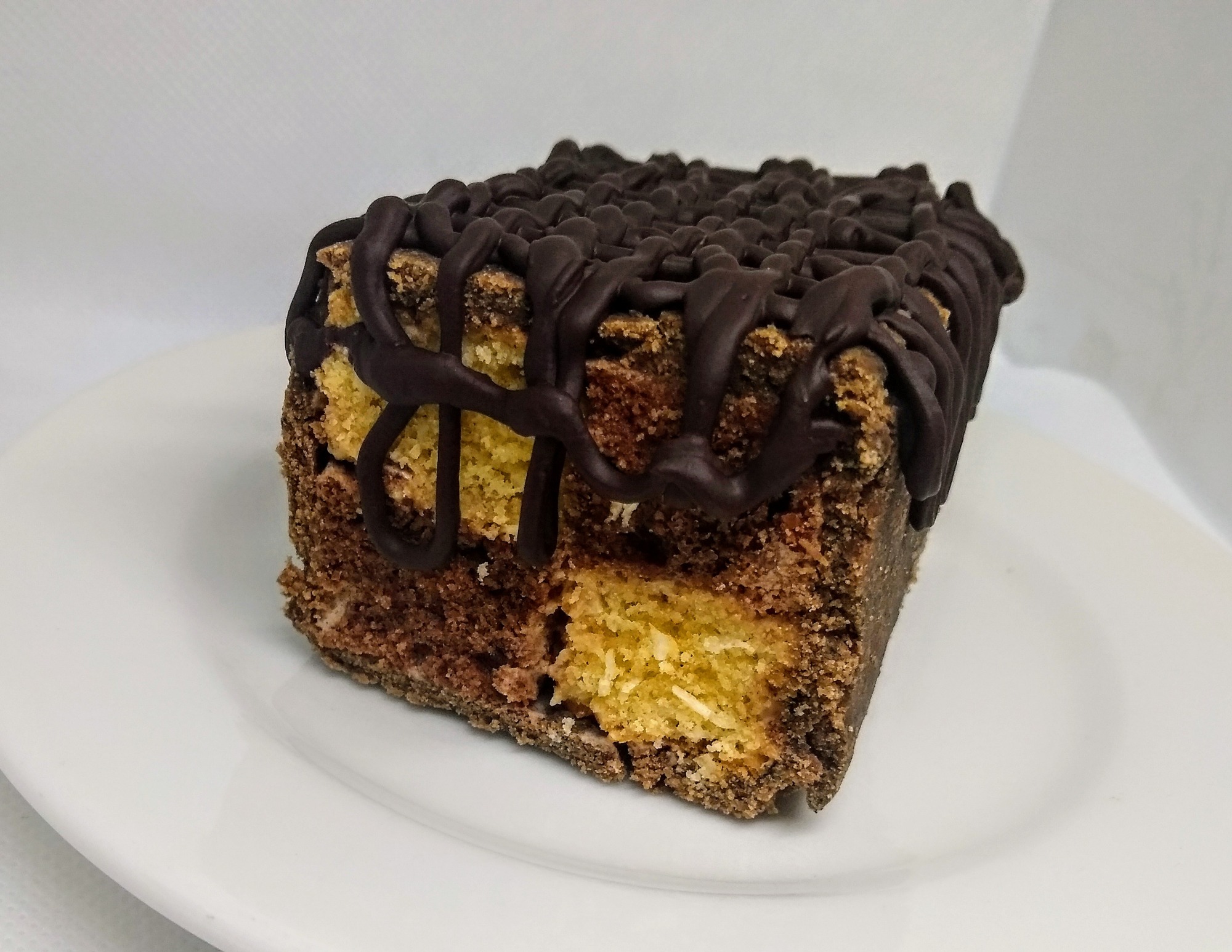 Kwadratowy kawałek ciasta z polewą czekoladową na białym talerzu, widoczne warstwy jasnego i ciemnego ciasta.