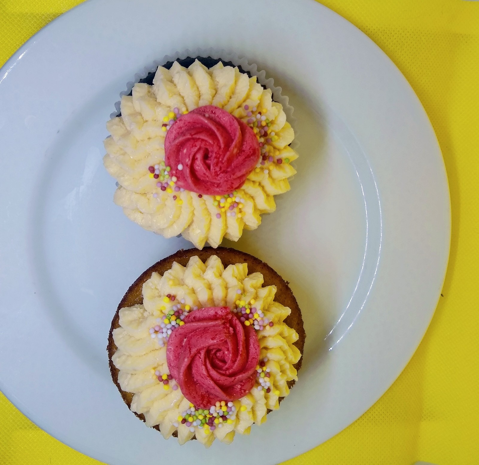 Dwa babeczki z kremem w kształcie kwiatów, ozdobione kolorowymi posypkami, ułożone na białym talerzu z żółtym tłem.