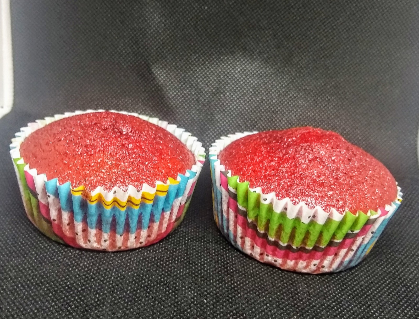 Dwa czerwone babeczki w kolorowych papilotkach na ciemnym tle.