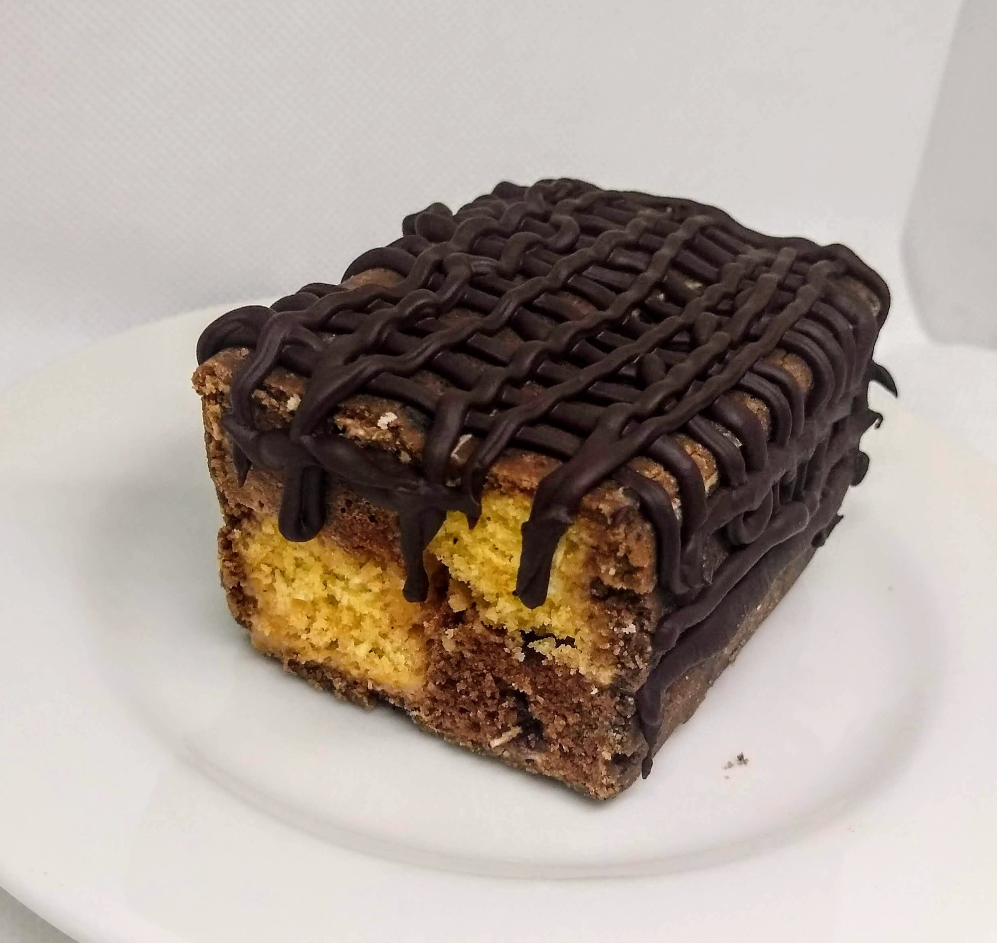 Kwadratowy kawałek ciasta z dwuwarstwowym biszkoptem, polany gęstą, ciemną polewą czekoladową, leżący na białym talerzu.