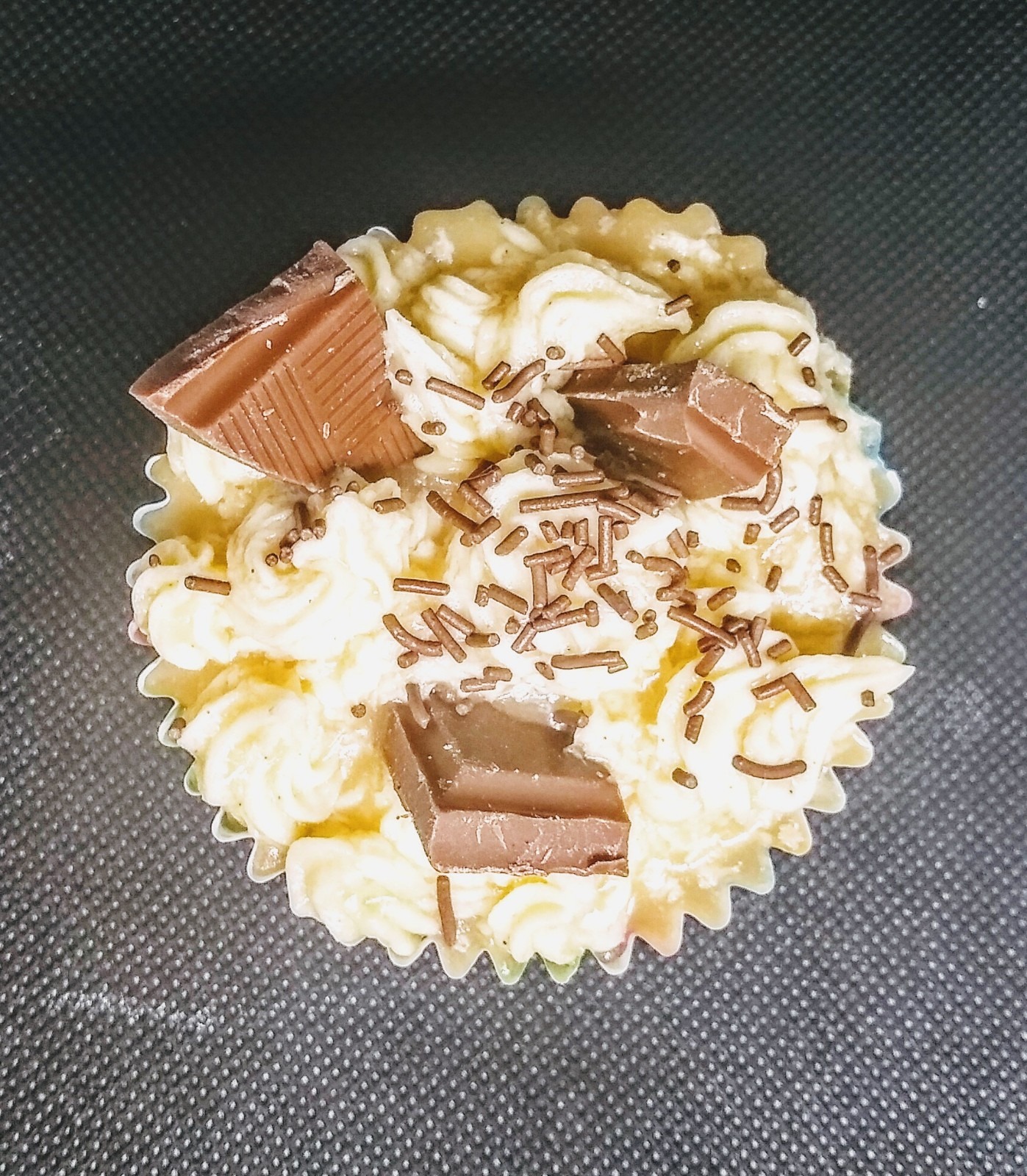 Kremowy babeczka z kawałkami czekolady i posypką czekoladową, umieszczona w papierowej foremce z falbankami, widok z góry na ciemnym tle.