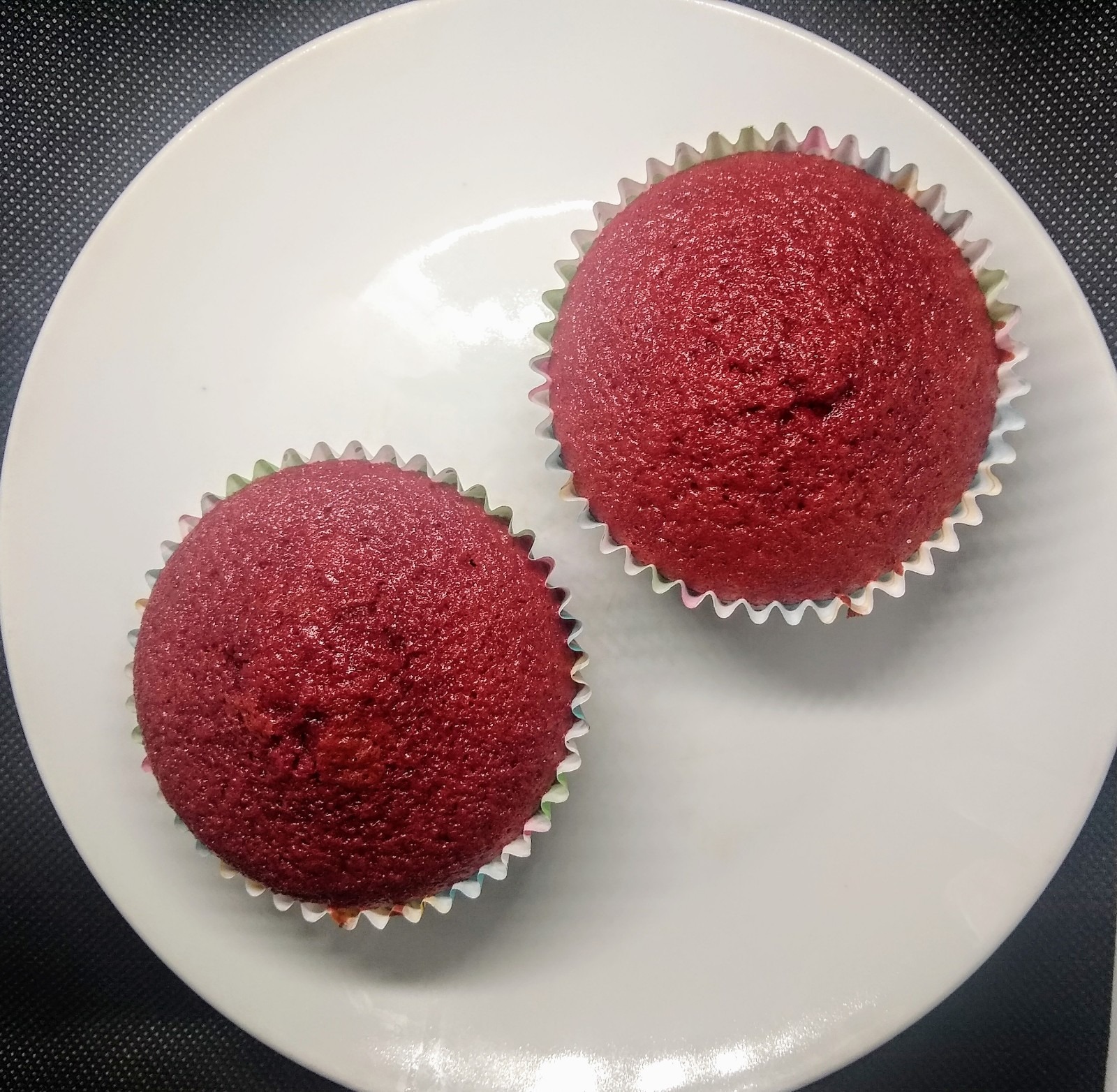 Dwa czerwone babeczki w papierowych foremkach, ułożone na białym talerzu na ciemnym tle.
