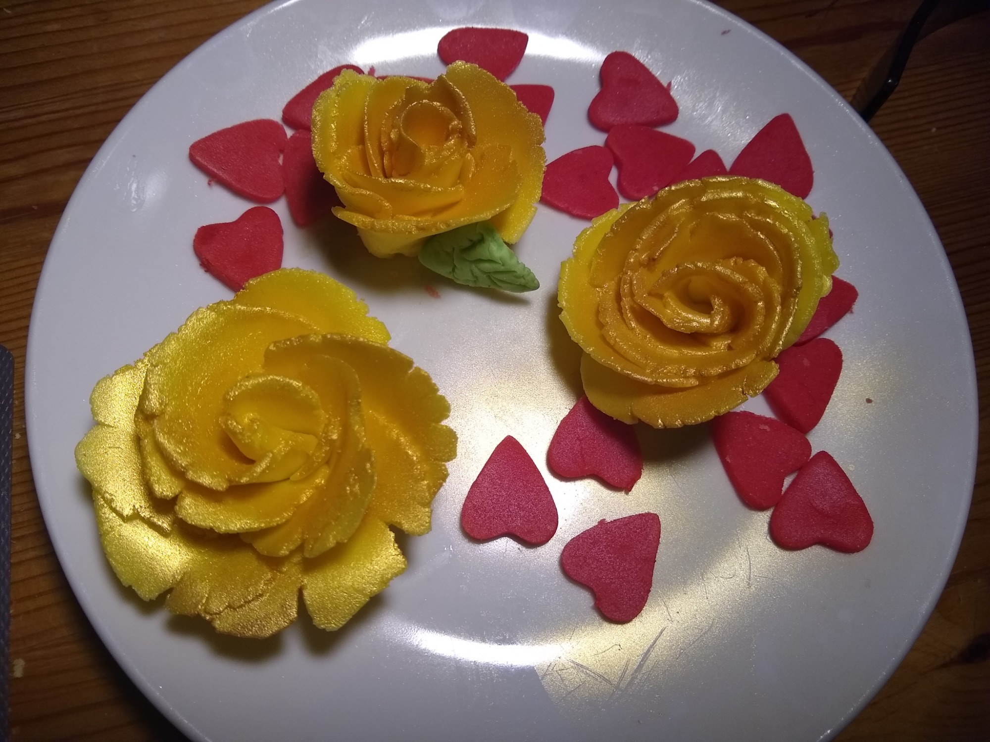 Trzy żółte, lukrowane róże z zielonym listkiem na białym talerzu, otoczone czerwonymi, cukrowymi sercami.