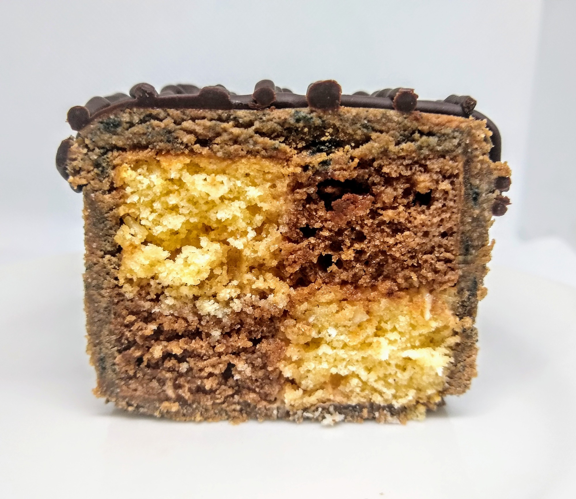 Przekrój kostki dwukolorowego ciasta marmurkowego z czekoladową polewą i dekoracją z małych kuleczek, na białym talerzu.