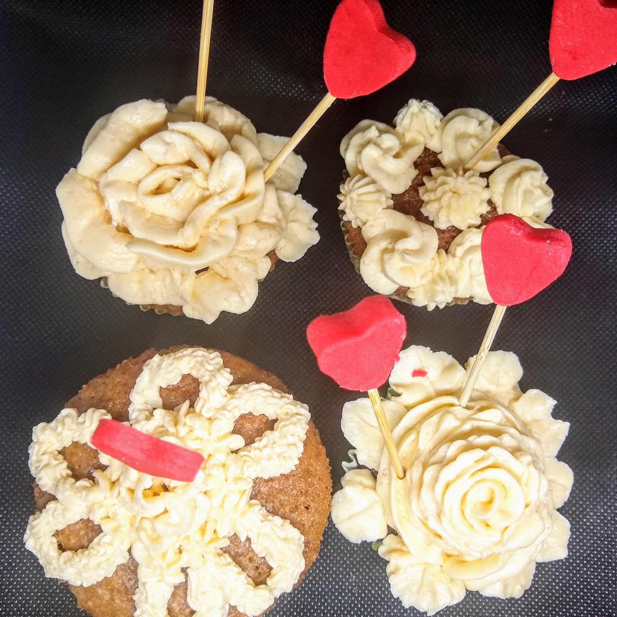 Cztery babeczki z kremem w kształcie kwiatów i czerwonymi serduszkami na patyczkach, ułożone na ciemnym tle z fakturą.