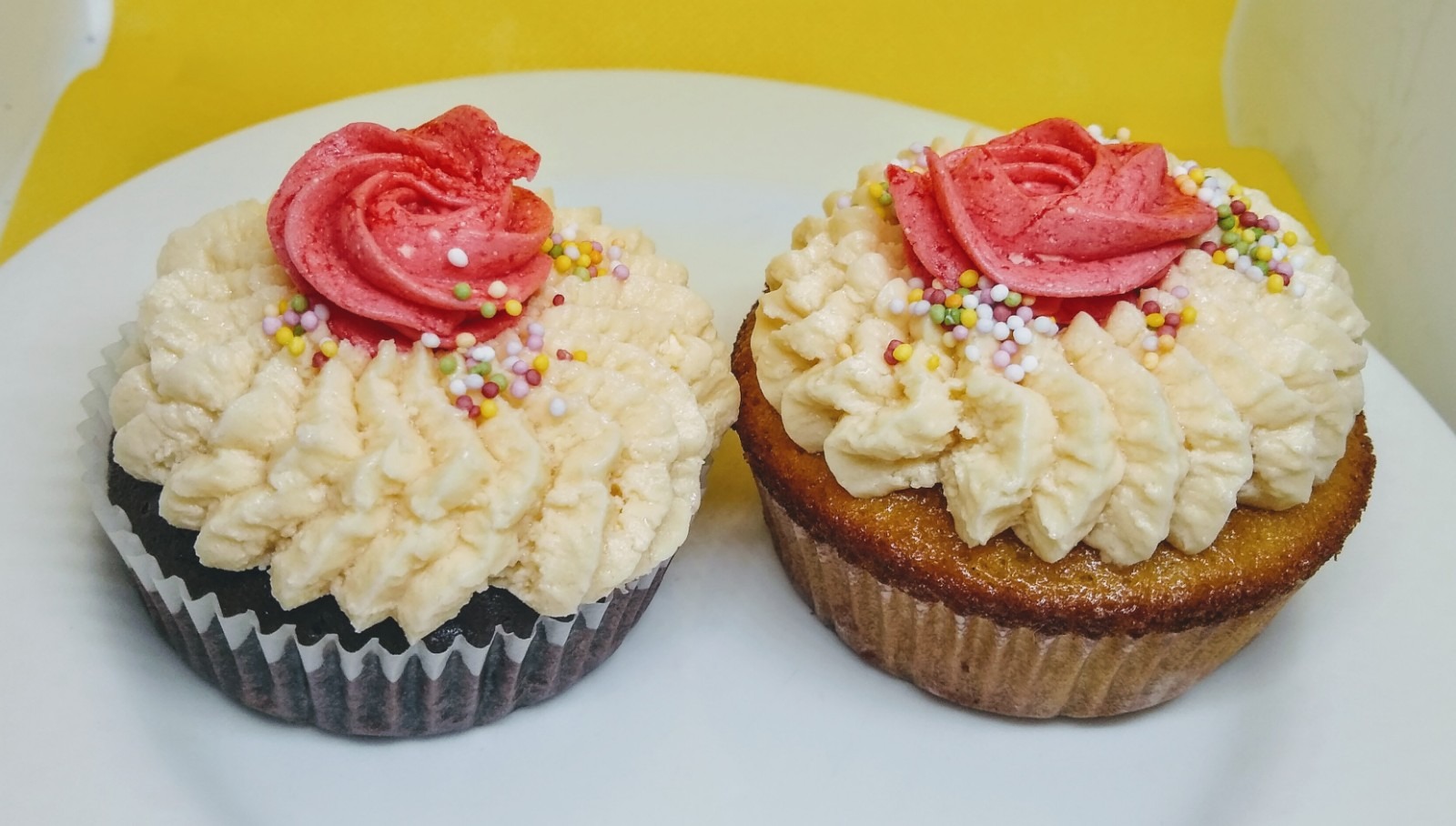 Dwa babeczki z kremem w odcieniach beżu i dekoracją z różowego kremu w kształcie róży, posypane kolorową posypką na białym talerzu.