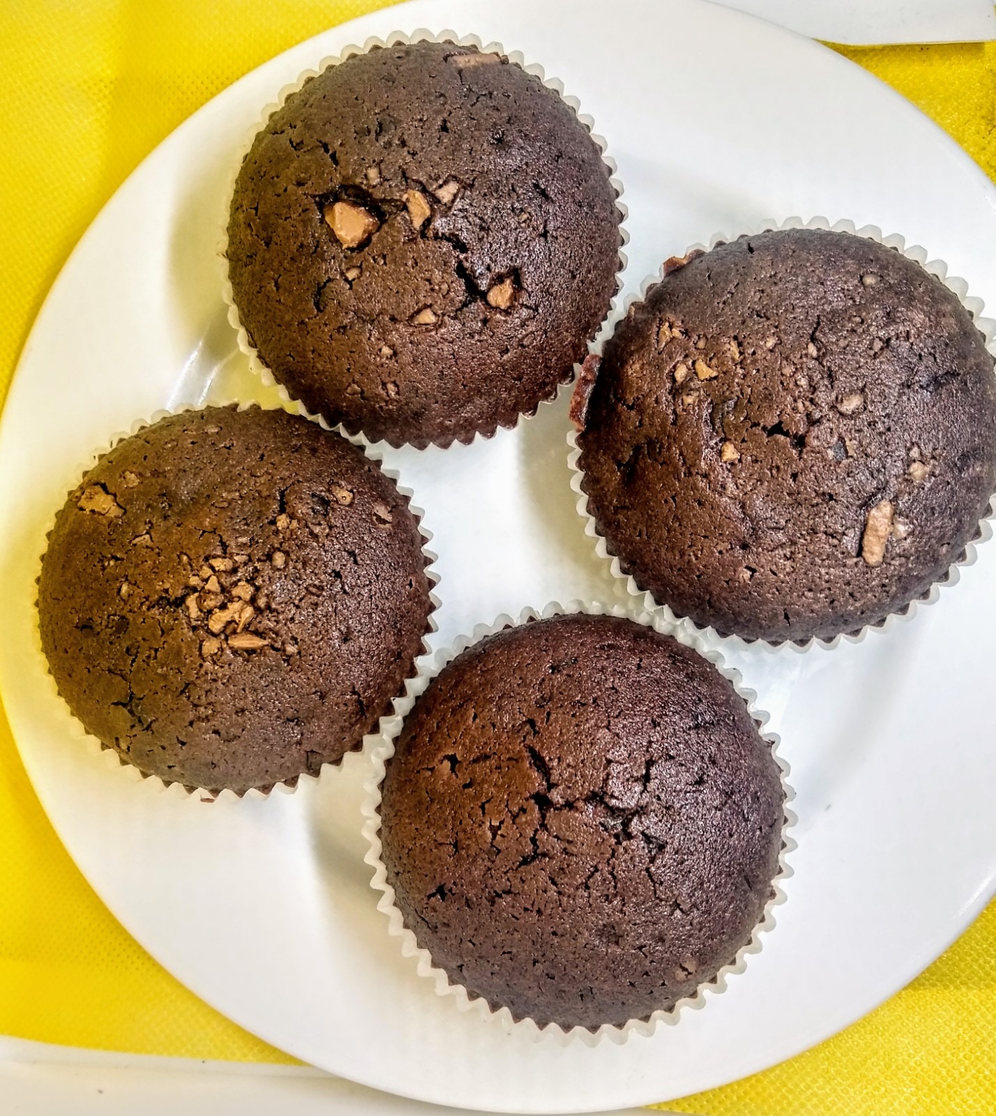 Cztery czekoladowe babeczki z kawałkami czekolady w papilotkach na białym talerzu, na żółtym tle.