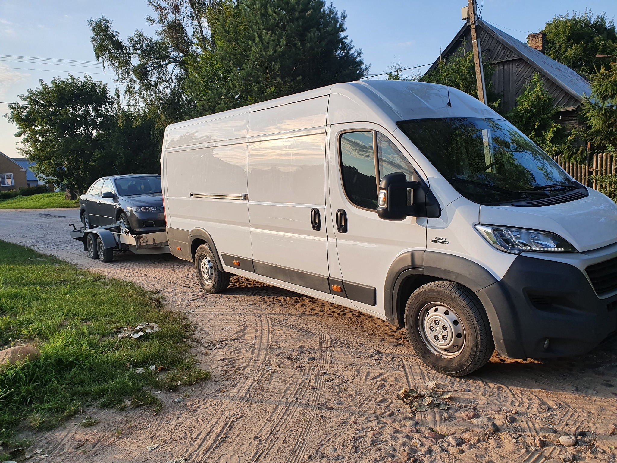 Biały bus z przyczepą przewożącą szary samochód osobowy na piaszczystej drodze wiejskiej, w tle drewniany budynek gospodarczy i drzewa.