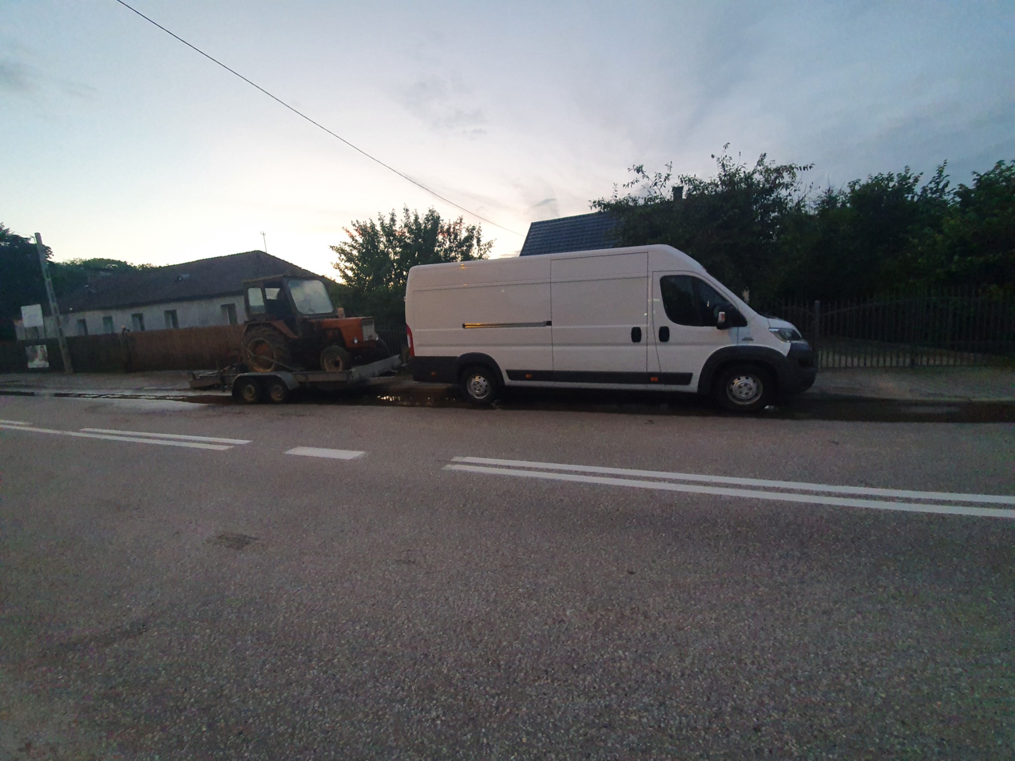 Biały bus zaparkowany na poboczu drogi, z przyczepą i traktorem na niej, na tle zabudowań i drzew przy zachmurzonym niebie.