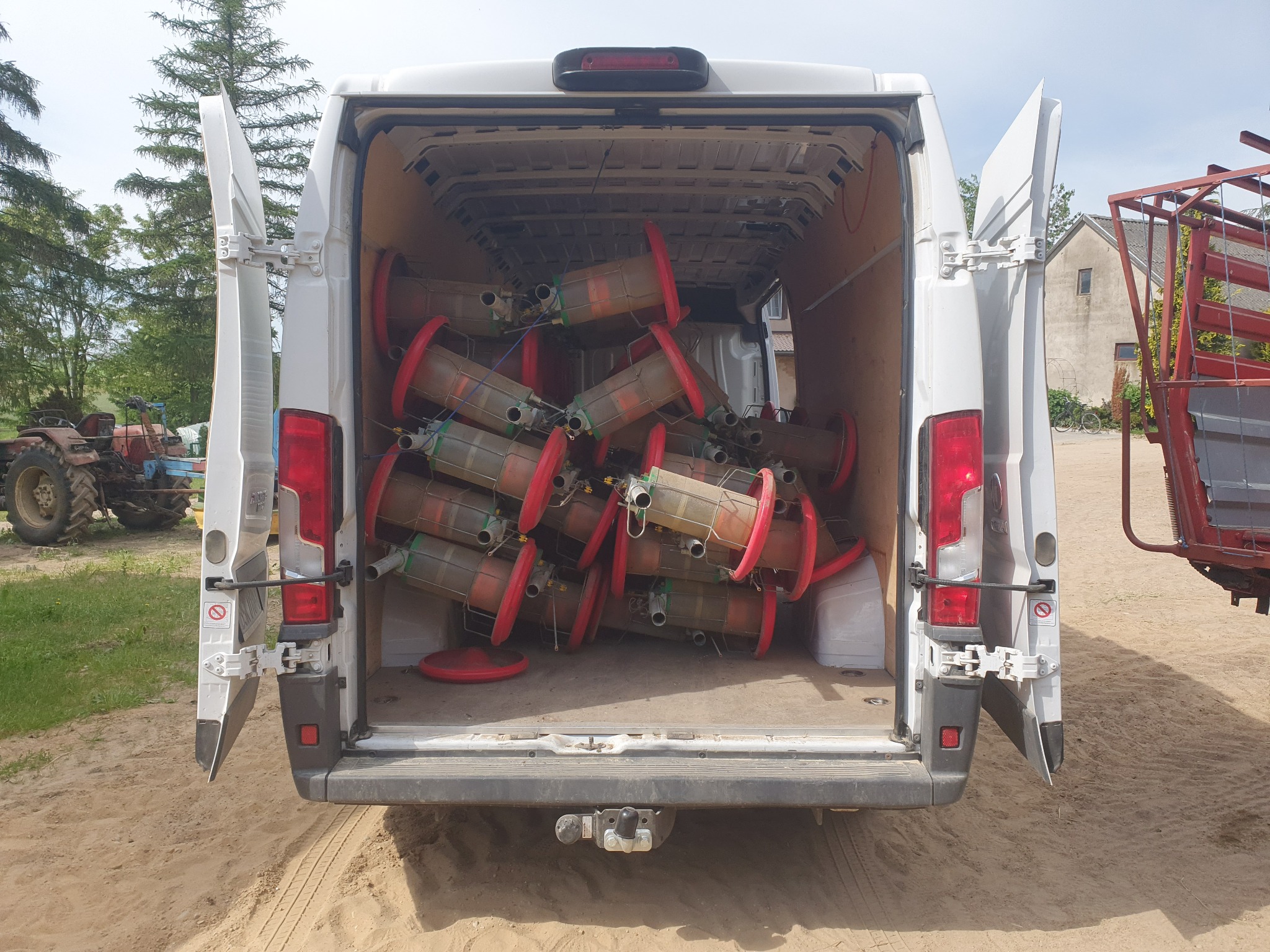 Wnętrze białego busa wypełnione cylindrycznymi pojemnikami z czerwonymi obręczami, widok z tyłu na otwarte drzwi, w tle traktor i budynek.