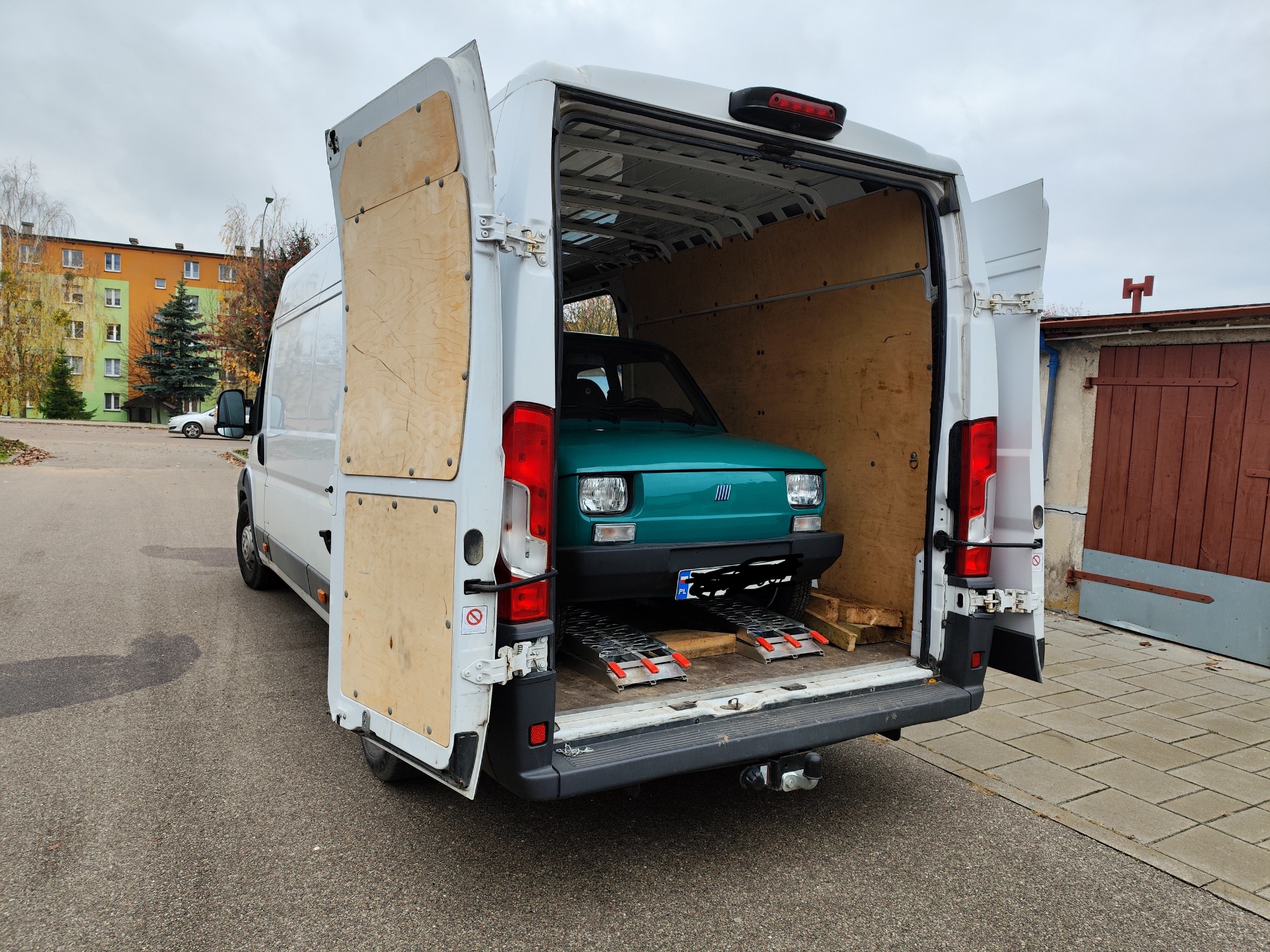Zielony Fiat 126p załadowany do białego busa z otwartymi drzwiami, widoczne rampy najazdowe.