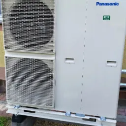 Zewnętrzna jednostka pompy ciepła Panasonic R32, zamontowana na betonowych blokach z podsypką z kamyków, na tle ściany budynku.