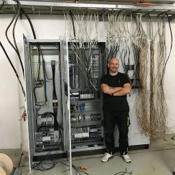 MODERN - TECH Technika domów - Uśmiechnięty elektryk z założonymi rękami stoi obok otwartej szafy sterowniczej z licznymi kablami, widoczne także drabina i zwinięte kable na podłodze.