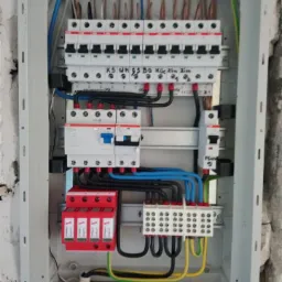 Otwarta skrzynka elektryczna z zainstalowanymi bezpiecznikami, przewodami i listwami zaciskowymi, widoczne ślady prac murarskich na ścianie.