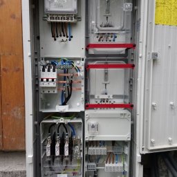 MODERN - TECH Technika domów - Otwarta skrzynka elektryczna z licznikiem, bezpiecznikami i różnymi złączami kablowymi, widoczne kable wchodzące do skrzynki od dołu.