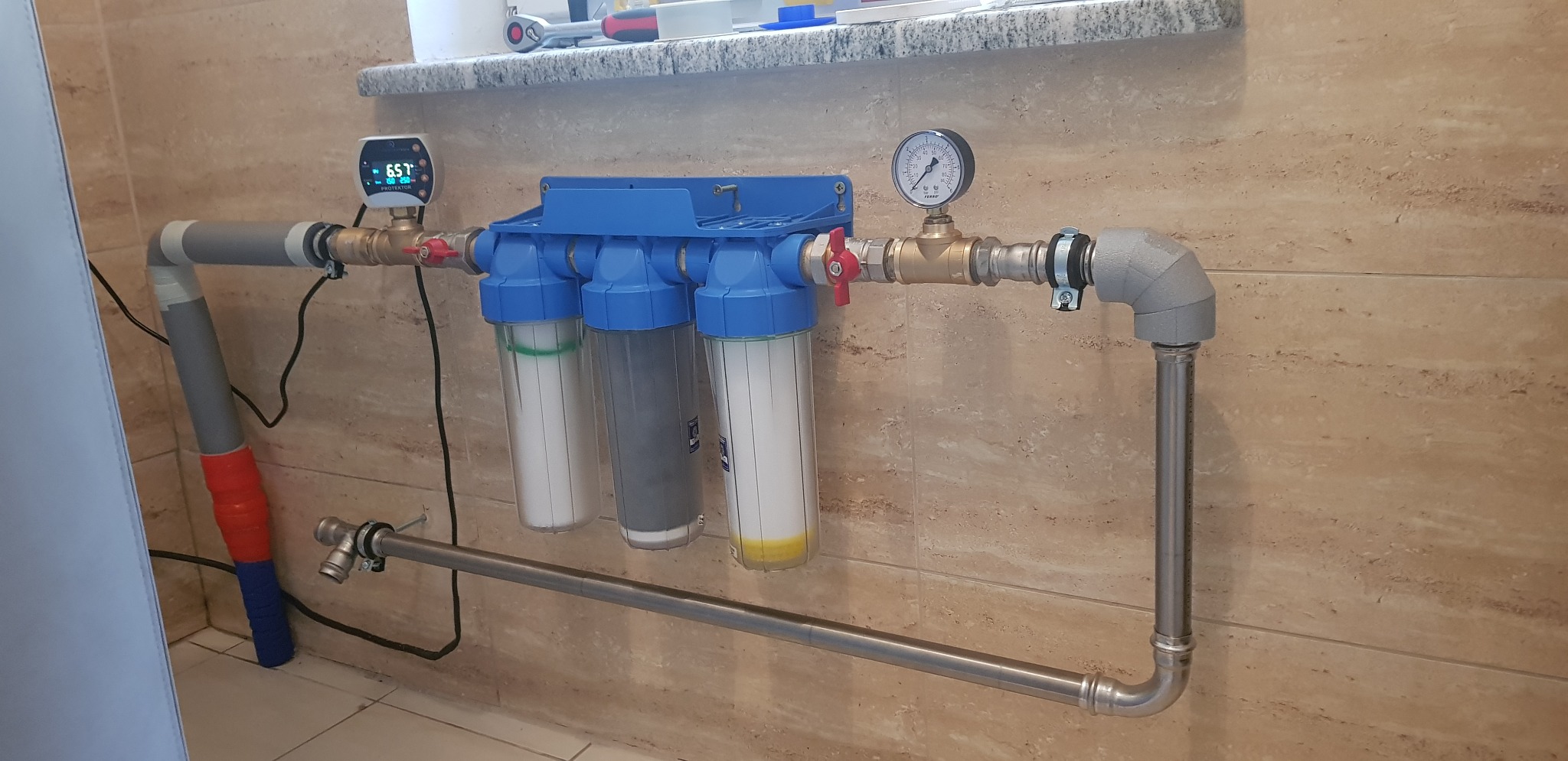 Zamontowany, niebieski system filtrów wody z trzema przezroczystymi obudowami, podłączony do rur, z manometrem i elektronicznym wyświetlaczem temperatury, na tle beżowej ściany z płytek.