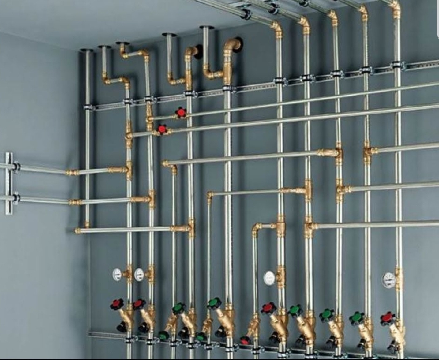 Rozbudowana instalacja rurowa z licznymi zaworami i manometrami, zamontowana na szarej ścianie, z widocznymi elementami mocującymi.