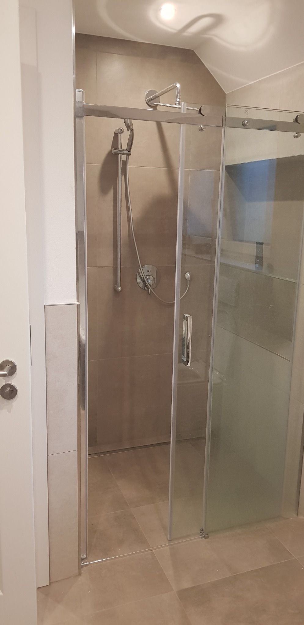 Zamknięta szklana kabina prysznicowa z przesuwnymi drzwiami, chromowane elementy, beżowe płytki ścienne i podłogowe, widoczna słuchawka prysznicowa i bateria.