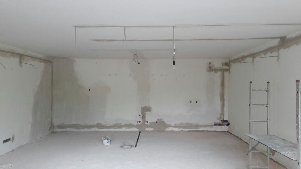 Wnętrze w trakcie remontu: surowe ściany z widocznymi punktami instalacji elektrycznej, wiszące przewody, drabina aluminiowa i wiadro z zaprawą na podłodze.