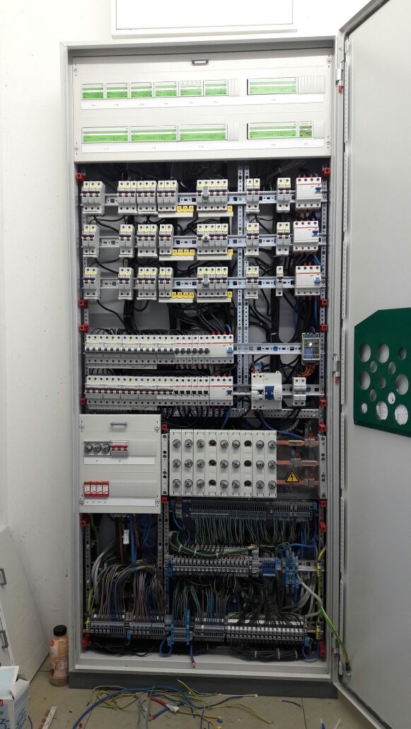 Otwarta szafa elektryczna z widocznymi zabezpieczeniami, bezpiecznikami, przewodami i listwami zaciskowymi; widoczny znak ostrzegawczy o wysokim napięciu; na podłodze leżą luźne przewody.