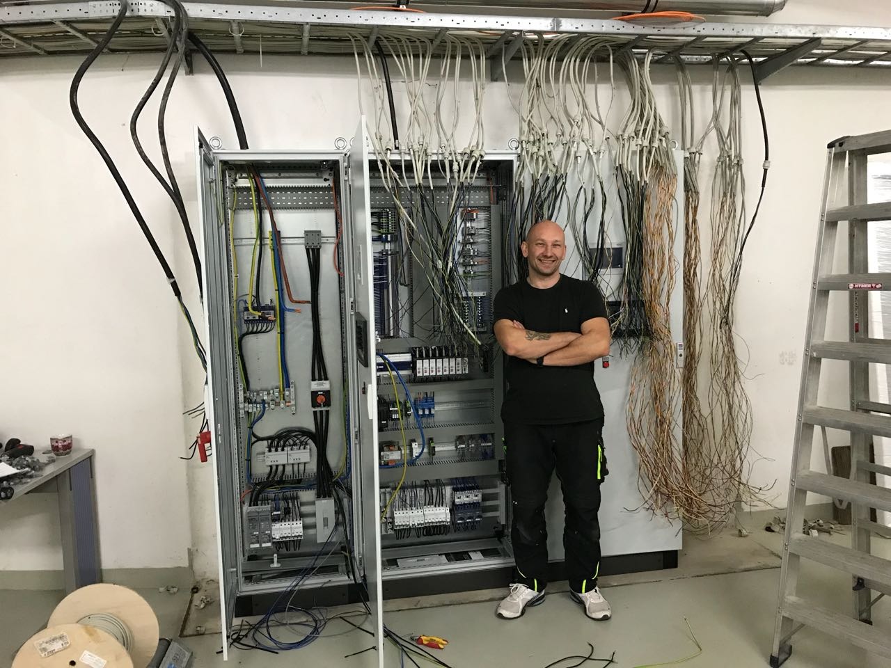Uśmiechnięty elektryk z założonymi rękami stoi obok otwartej szafy sterowniczej z licznymi kablami, widoczne także drabina i zwinięte kable na podłodze.